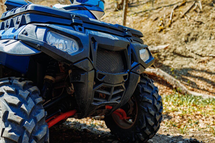 Close-up view of a rugged blue ATV parked on a forest trail with visible off-road tires and headlights.